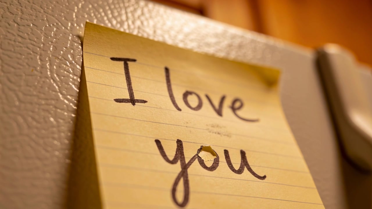 La telecamera si avvicina mostrando un post-it giallo con scritto 'Ti amo' sul frigorifero della cucina, rivelando una lacrima.