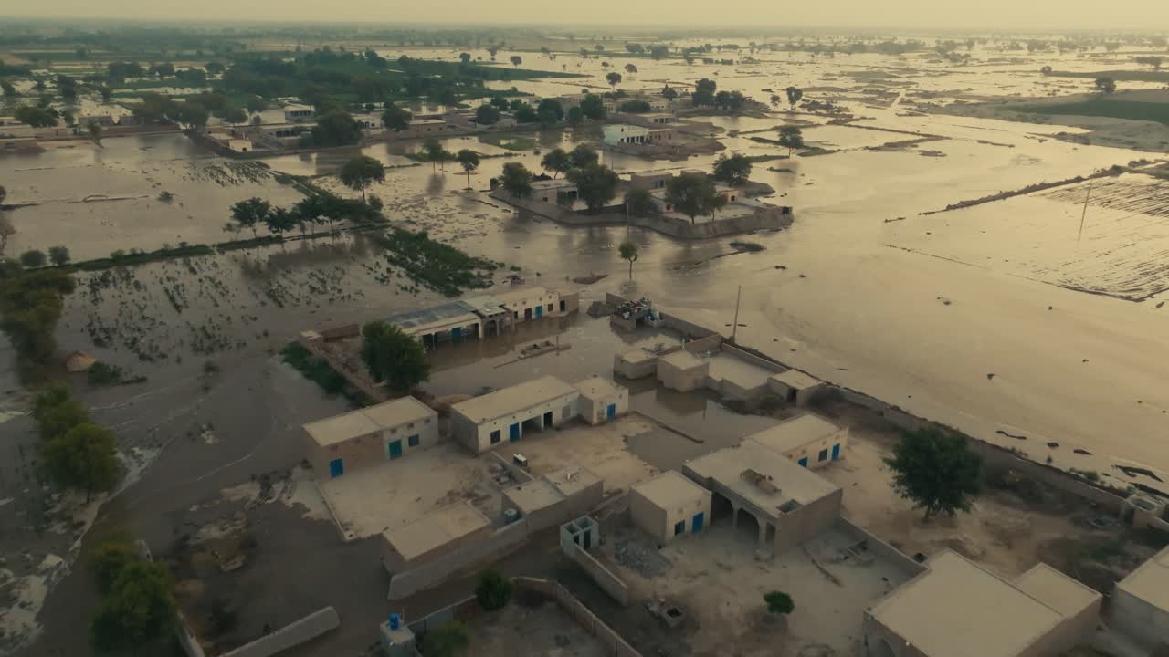 Drone overpasses a large residential complex surrounded by flooded fields during the afternoon in Pakistan, with white birds going by