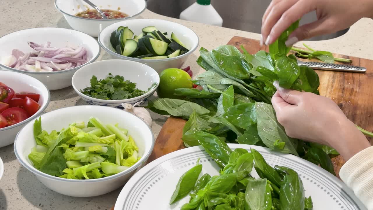 A person’s hands pick and prepare fresh Thai basil leaves on a wooden cutting board, surrounded by bowls of colorful vegetables in bright kitchen lighting