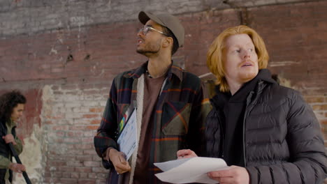 two production coworkers talking and reading a document about the movie in a ruined building while other colleagues talking and waiting to start the recording