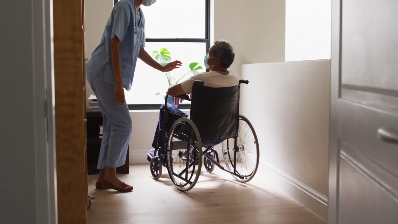 médico de raza mixta con máscara asistiendo a un anciano en silla de ruedas en casa