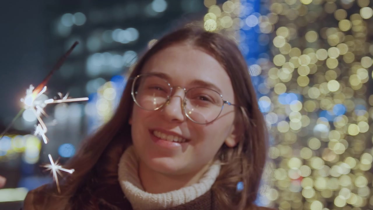 Stadtlichter funkeln. Frau hält leuchtendes Feuerwerk. Nachtszene mit leuchtender Wunderkerze in der Hand. Nächtliche Stadtlandschaft mit einer Frau, die ein strahlendes, funkelndes Feuerwerk in der Hand hält.