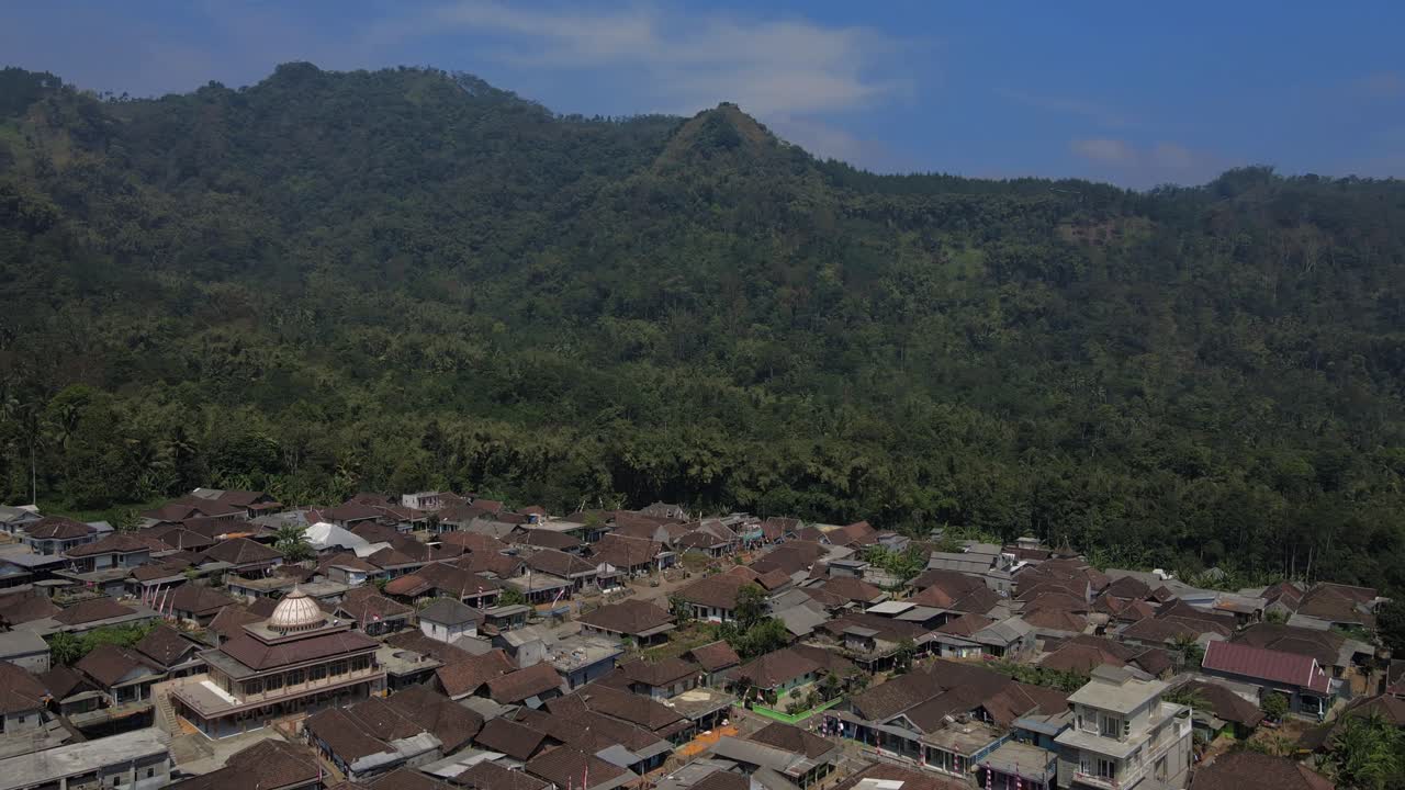 Pemandangan dari drone udara desa di Indonesia dengan masjid dan atap tradisional, berlatar belakang perbukitan tropis yang rimbun dan langit biru yang cerah