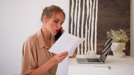 Blonde-Frau-Bespricht-Papierkram-Am-Telefon-In-Der-Nähe-Des-Laptops