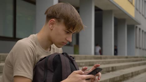 youngster sitting in front of the school and scrolling mobile phone with his backpack in front