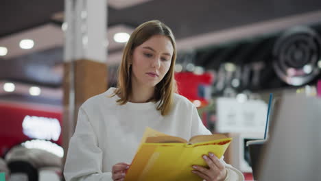 young lady reading yellow book while seated indoors in mall environment with soft blurred background, thoughtful expression showing focus and engagement