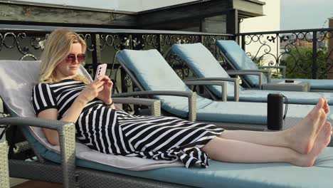 blonde fashion girl vibing to music lying on a rooftop deck chair at day time with bluetooth speaker next to her