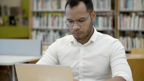 thoughtful young man typing on laptop in library