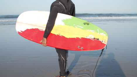 surfista masculino con una pierna artificial parada en la orilla del océano y sosteniendo una tabla de surf bajo el brazo