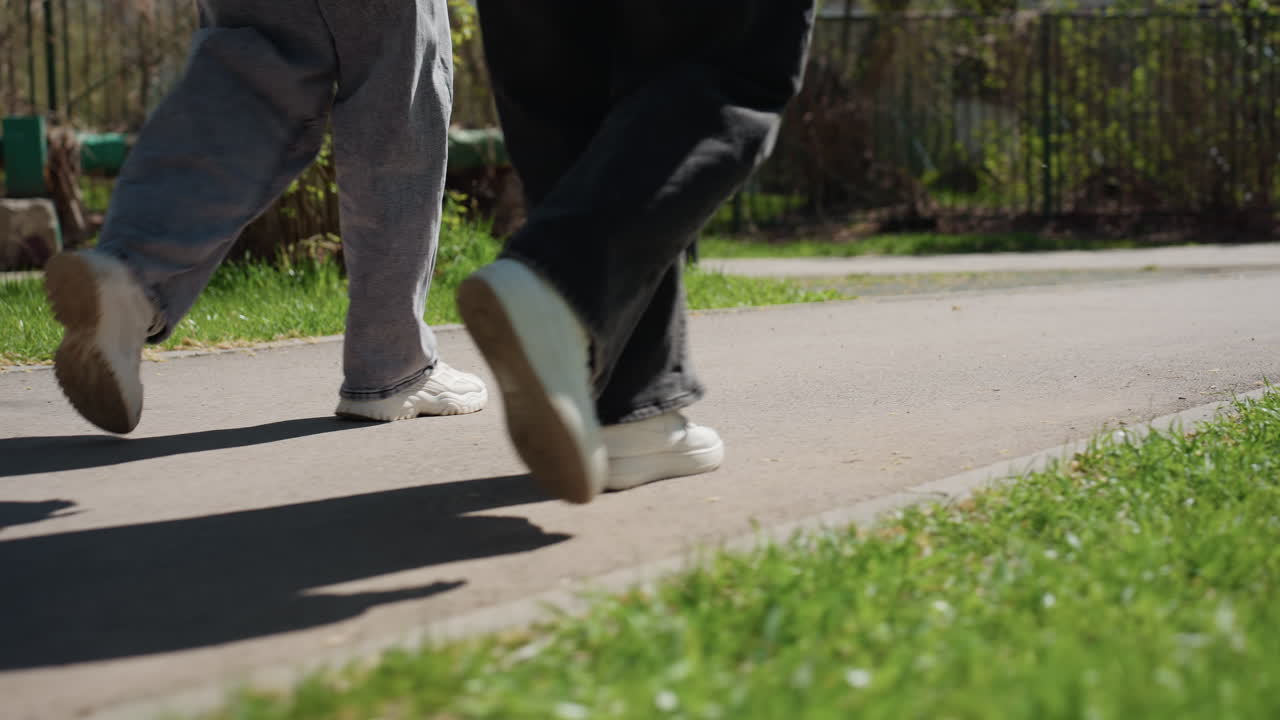 公園の小道を並んで歩く2人、足音と長い影、カジュアルなスニーカーとリラックスしたペース、緑の芝生と太陽光、静かな仲間意識とリズミカルな動きを後方から捉えた写真。
