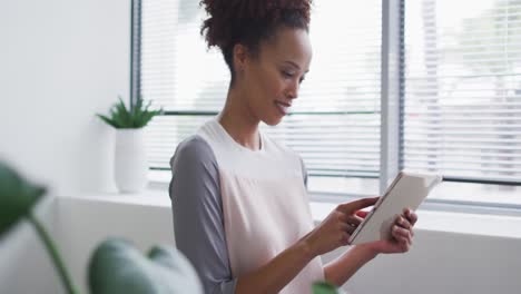 Smiling-mixed-race-businesswoman-using-digital-tablet-and-standing-by-window-in-office