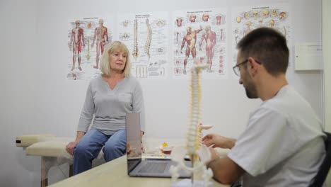 female patient talking to doctor about her problems with her back during consultation in the doctor female patient talking to doctor about her problems with her back during consultation in the doctors office physiotherapist showing spine model to his patient in medical office