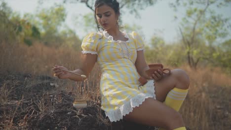 wide shot of young beautiful indian woman dipping a brush into a pot resting on the ground while fixing a broken clay pot