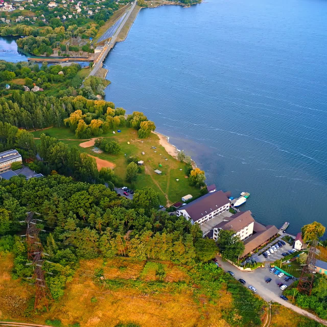Hotel located on the river bank with few piers and boats. Green waterfront and dam at backdrop. Aerial view