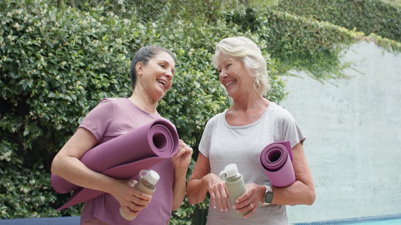 Ältere Freundinnen plaudern und kühlen sich nach dem Pool-Yoga ab, wobei sie lila Matten und Flaschen halten.