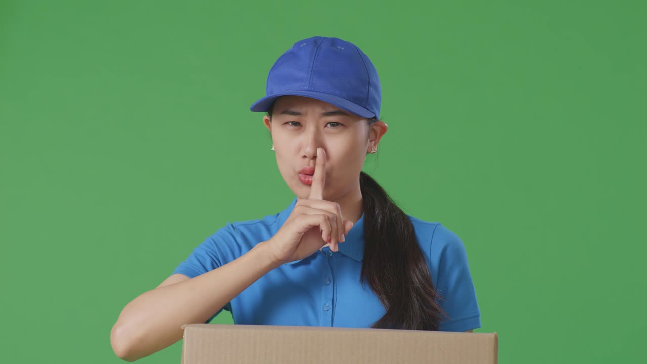 푸른 유니폼을 입은 아시아 여성 택배사의 클로즈업은 침 제스처를 보여주고 스튜디오에서 녹색 화면 배경에 상자를 전달하는 동안 머리를 흔들고 있습니다.