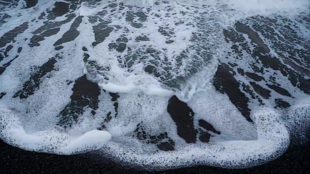 Arriving swell, white surf foam advancing and spreading across dark pebble beach, leaving channels