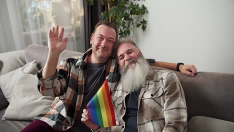 retrato de un hombre feliz con cabello gris y una barba blanca exuberante en una camisa a cuadros que sostiene una bandera lgbt junto con su novio moreno en una camiseta a cuadros sentado en un sofá gris en un apartamento moderno por la noche