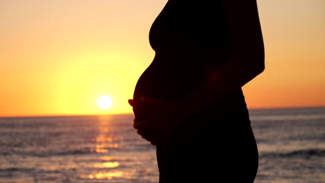 close up silhouette of pregnant woman touching belly in front of tranquil ocean during golden sunset light