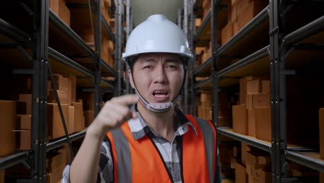 primer plano de un ingeniero masculino asiático con casco de seguridad de pie en el almacén con estantes llenos de mercancías de entrega. estando enojado, señalándote y gritando en el almacenamiento