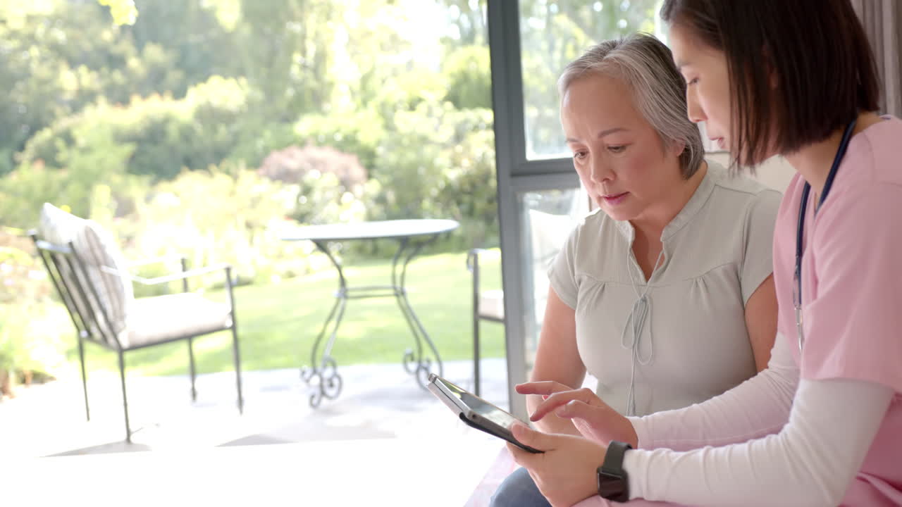 Physiotherapist showing senior asian woman exercises on tablet during home visit, copy space
