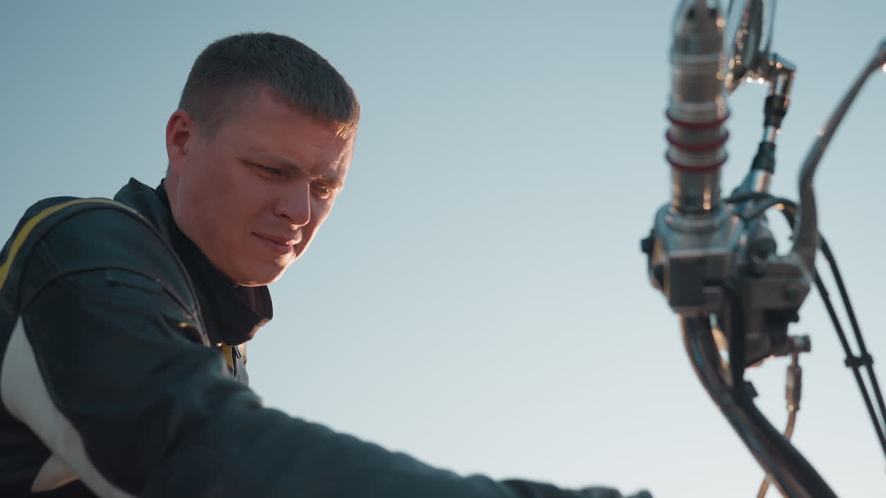 Biker in leather jacket carefully wipes motorcycle handlebar and chrome tank with yellow cloth, sunlight highlighting details of metal and leather in focused outdoor maintenance scene