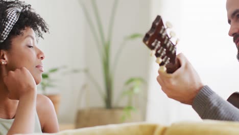 video de vista trasera de una feliz pareja diversa en casa, un hombre tocando la guitarra a una mujer, copia el espacio