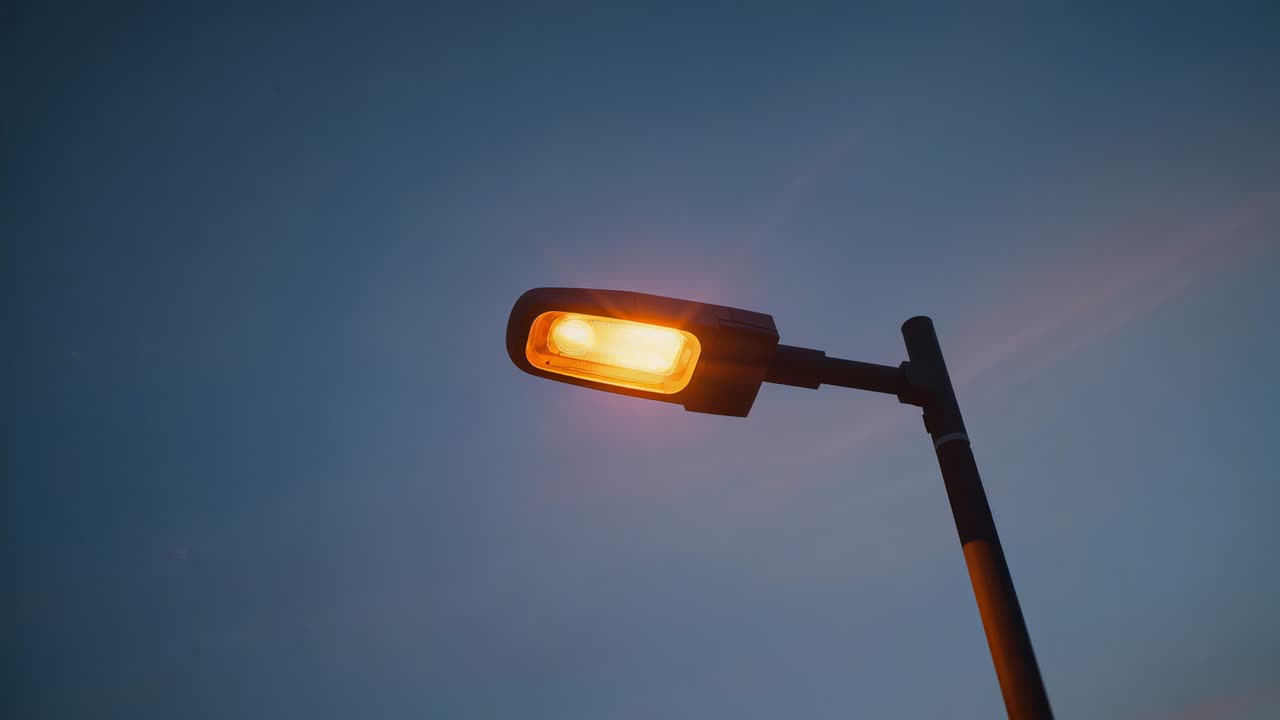 Lampu Jalan Bercahaya Memancarkan Cahaya Oranye Hangat di Tepi Jalan, dengan Kepala Lampu, Lengan, Tiang, Tersedia Ruang Teks