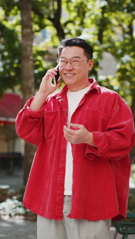 Happy-Asian-man-having-remote-conversation-talking-on-smartphone,-good-news-gossip-in-city-street