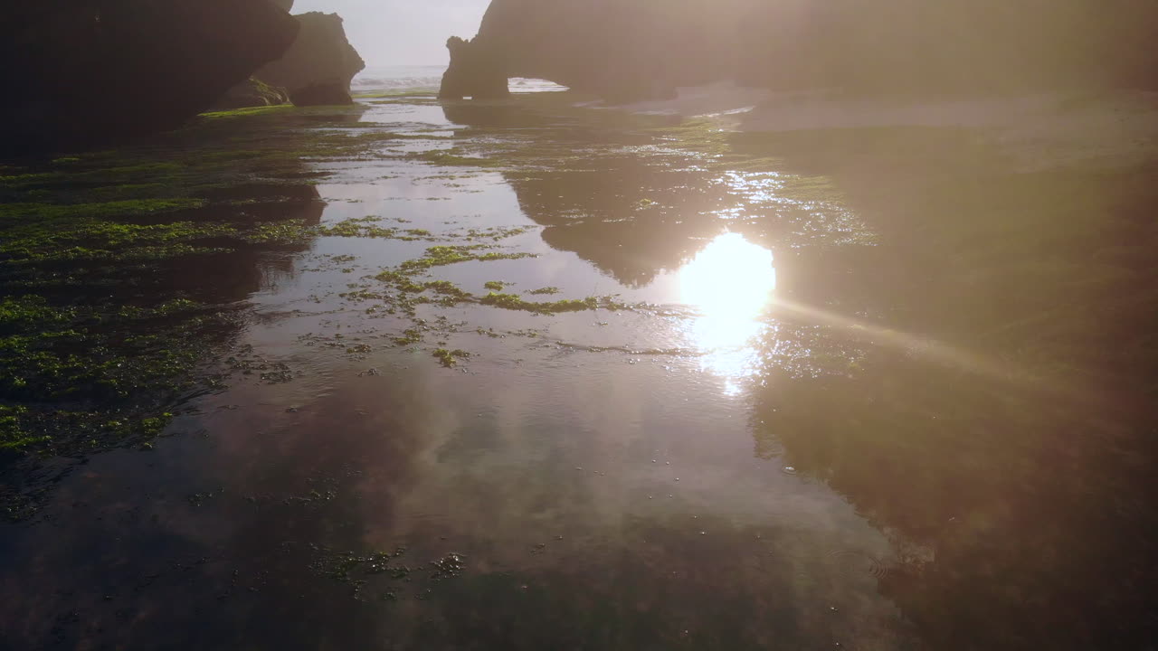 una toma mágica: el sol naciente se refleja en el océano en calma, una hermosa costa rocosa cubierta de algas, una gran formación rocosa en el fondo
