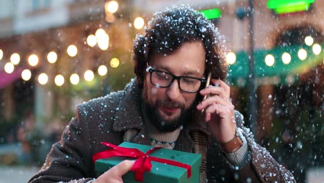 close-up view of caucasian man wearing eyeglasses holding a present and talking on smartphone on the street while it‚äôs snowing in christmas