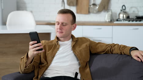 young guy writing messages on a phone at home. portrait of a handsome man sitting on a couch with a smartphone in the kitchen.