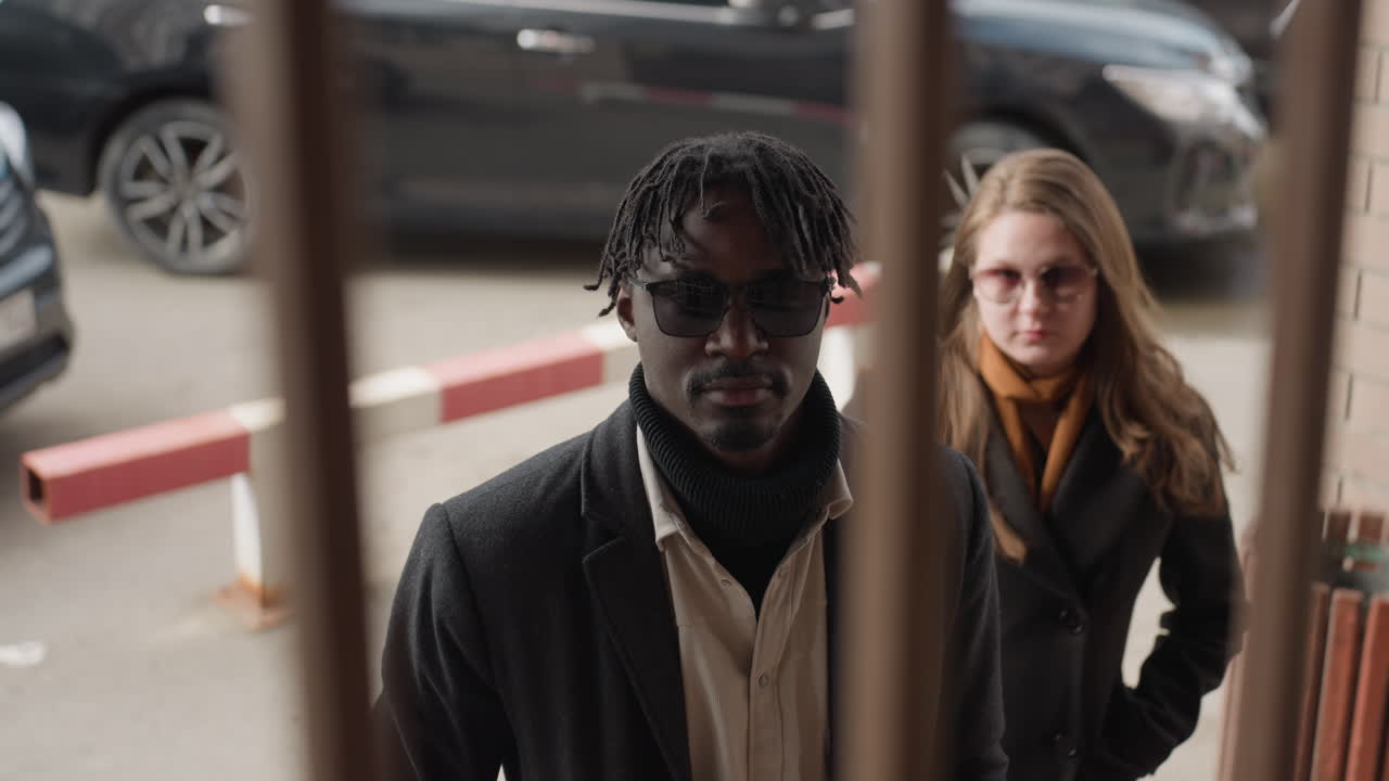 На фотографии мужчина и женщина в солнцезащитных очках за железной решеткой, стоящие возле парковочного барьера, спокойная поза при приглушенном дневном свете, неглубокая резкость и легкий ветерок, развевающий волосы.