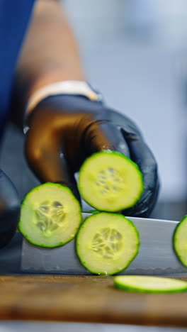 Unrecognized cook's hands in latex gloves cut the cucumber with a big sharp knife. Fresh vegetable recipe. Close up. Vertical video.