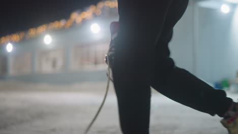side view of skater walking while holding black ice skating boot by lace near snow-covered ground at night, with festive building lights glowing in soft blur background