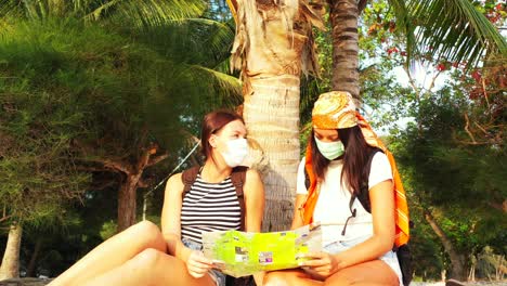 young women with face masks protecting from coronavirus reading a tourist guide of tropical island with paradise beaches in thailand