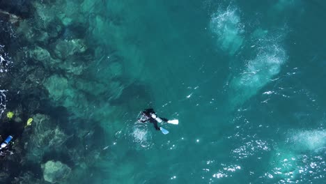 High-view-of-scuba-divers-exploring-a-rocky-shoreline-with-bubbles-breaking-the-ocean-surface