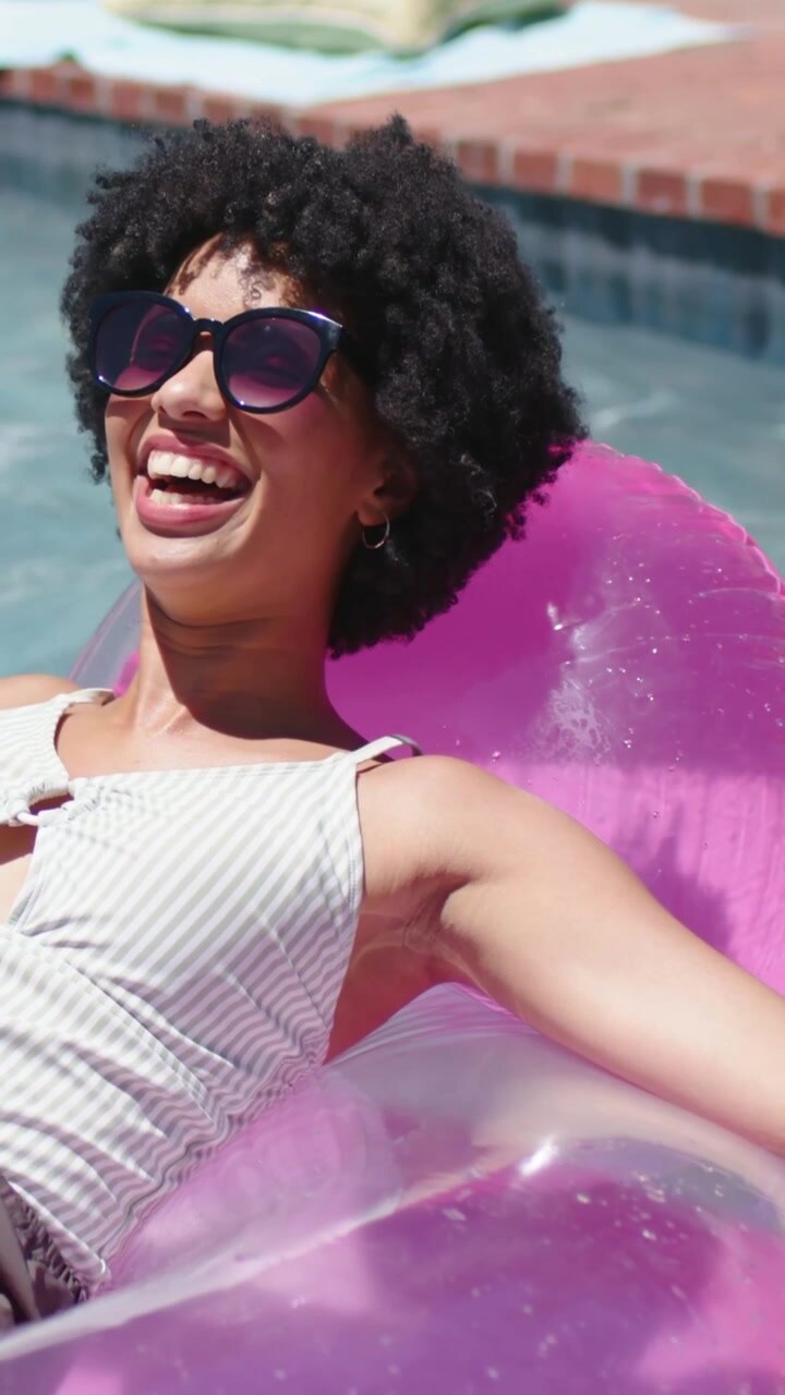 Vertical video: Cam zooming woman turning head laughing on pink float at pool, swim top and shades