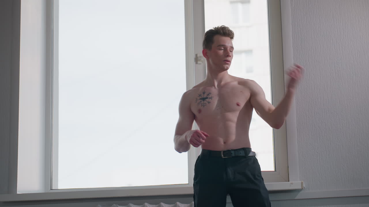 shirtless boy stands in front of window performing arm flexibility movement, relaxed posture with light streaming in, displaying toned upper body and tattoo while engaging in light fitness activity