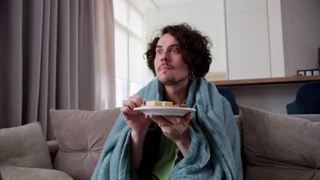 Interested-brunette-guy-with-curly-hair-wrapped-in-a-blue-blanket-and-watching-a-movie-while-eating-sweet-snacks-at-home-in-a-modern-apartment