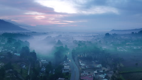 Breite-Luftaufnahmen-Der-Straße-Und-Autos,-Die-Im-Blauen-Licht-Des-Sonnenaufgangs-Der-Stadt-San-Juan-Ostuncalco-Und-Der-Dahinter-Liegenden-Berge-Im-Dunst-Fahren