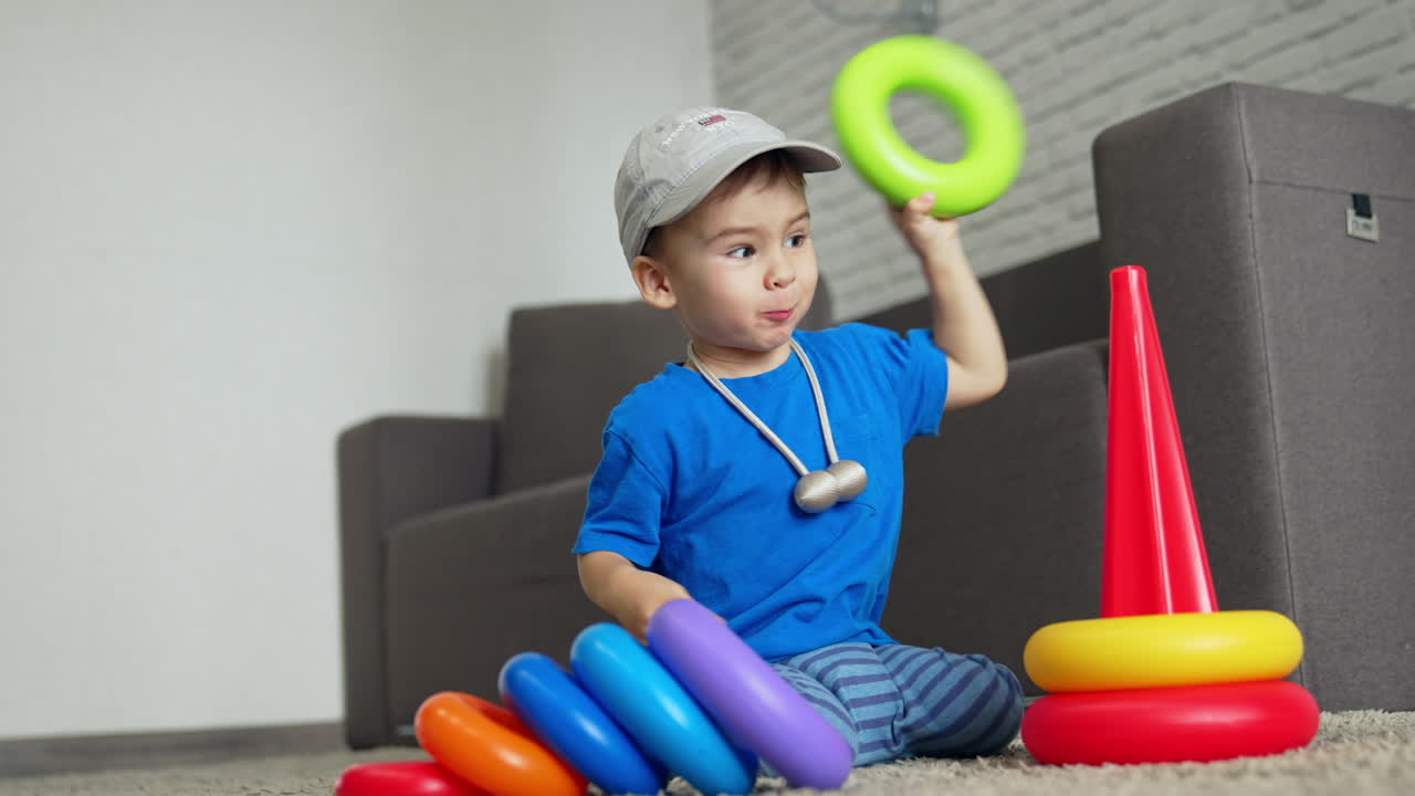 Lovely baby boy in a cap and blue t-shirt playing at home. Toddler collects a pyramid and pushes it to fall then. Low angle view.