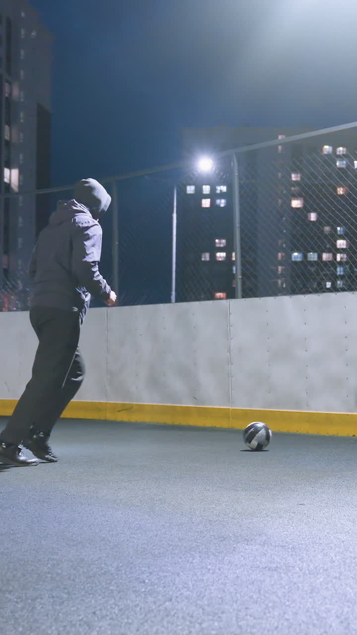 hombre pateando una pelota de fútbol contra la pared en un campo deportivo iluminado, mostrando la recreación nocturna urbana, el fondo incluye edificios altos con ventanas brillantes e iluminación vibrante