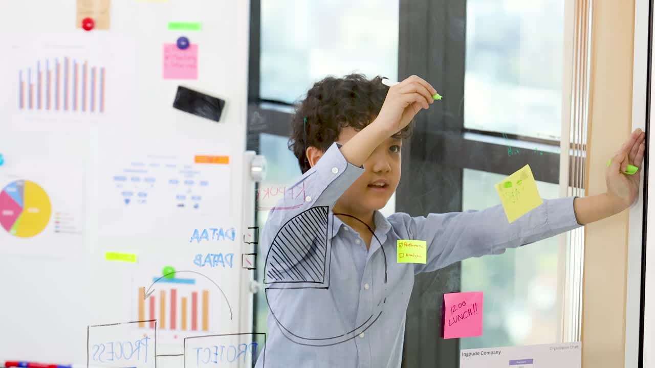 A young boy in a blue shirt draws data charts on glass in bright office