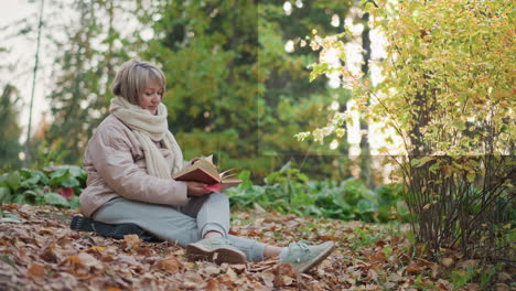 woman seated on forest ground flipping through book pages while surrounded by fallen leaves, golden autumn shrubs, and soft green foliage in quiet natural setting under warm afternoon sunlight
