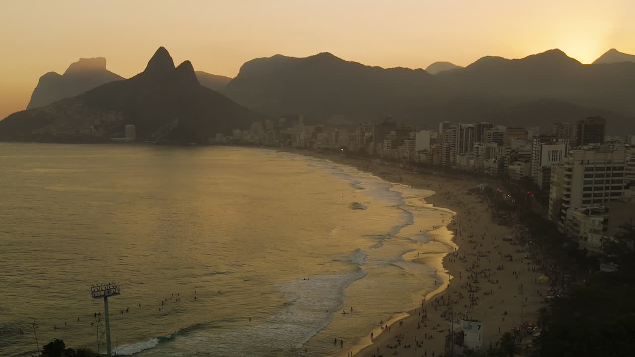 Drone descending over Ipanema Beach Rio de Janeiro Brazil at sunset, ocean reflection, skyscrapers, mountains background