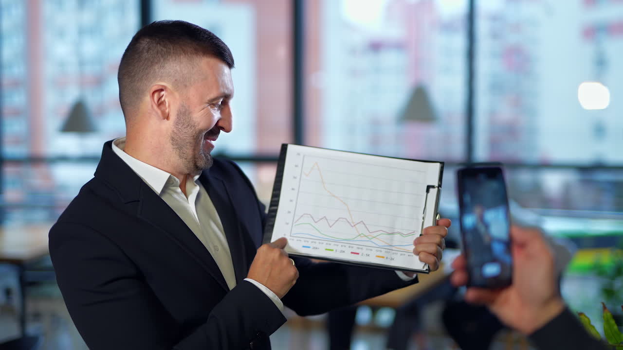 Hand with a phone taking video of a man with chart. Handsome man showing graph patterns and explaining them to the camera.