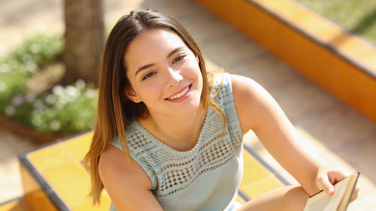 Mujer sonriente con blusa azul sosteniendo un libro en escalones amarillos, la luz del sol dirigiendo su mirada hacia arriba, mirando a la cámara.