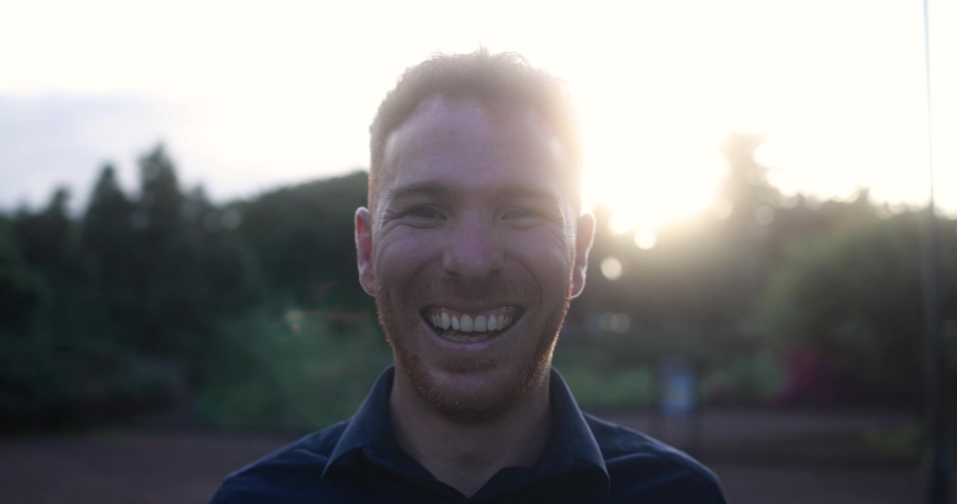 Uomo felice che sorride alla fotocamera con la luce del sole in controluce all'aperto