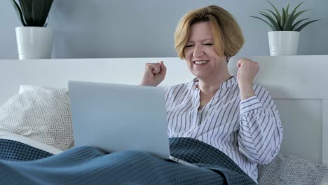 reaction of success by happy senior woman using laptop in bed